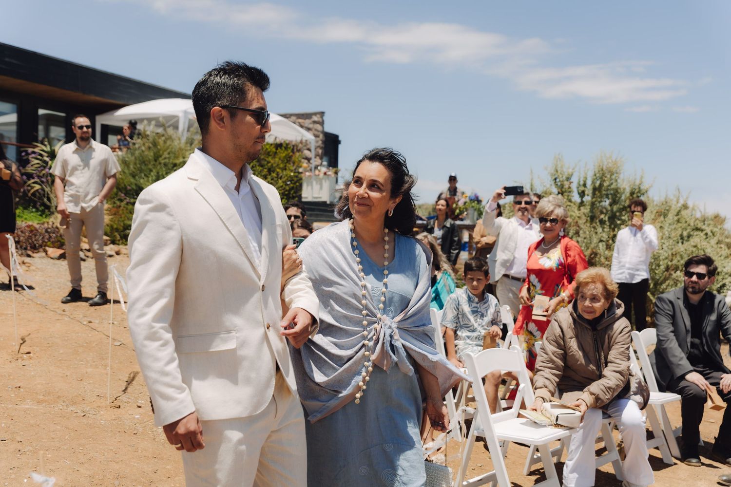 Smiling groom in cream suit walking with elegantly dressed woman in blue at outdoor wedding ceremony.