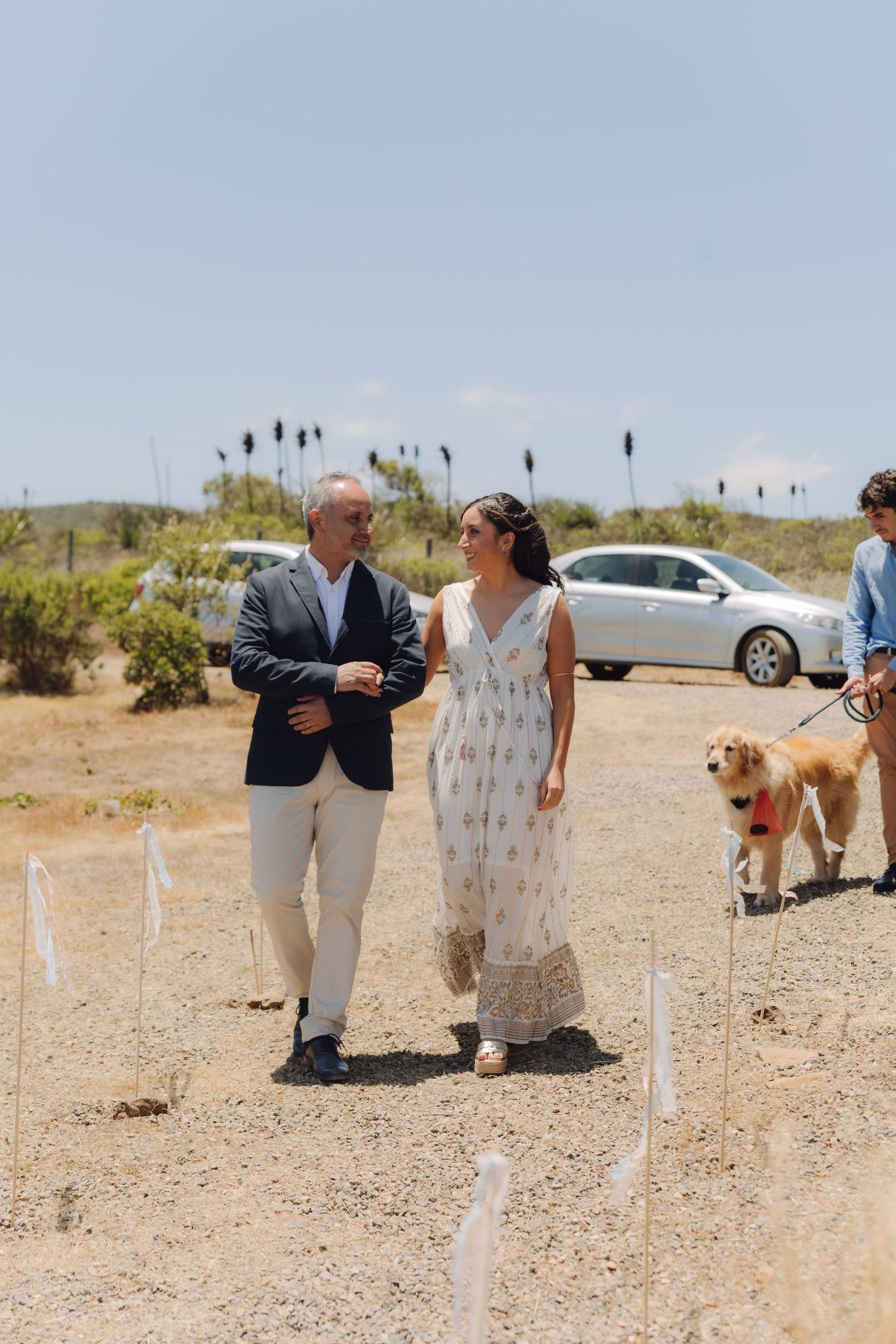 Father in navy blazer walking bride in floral dress down sandy outdoor wedding aisle with golden retriever.