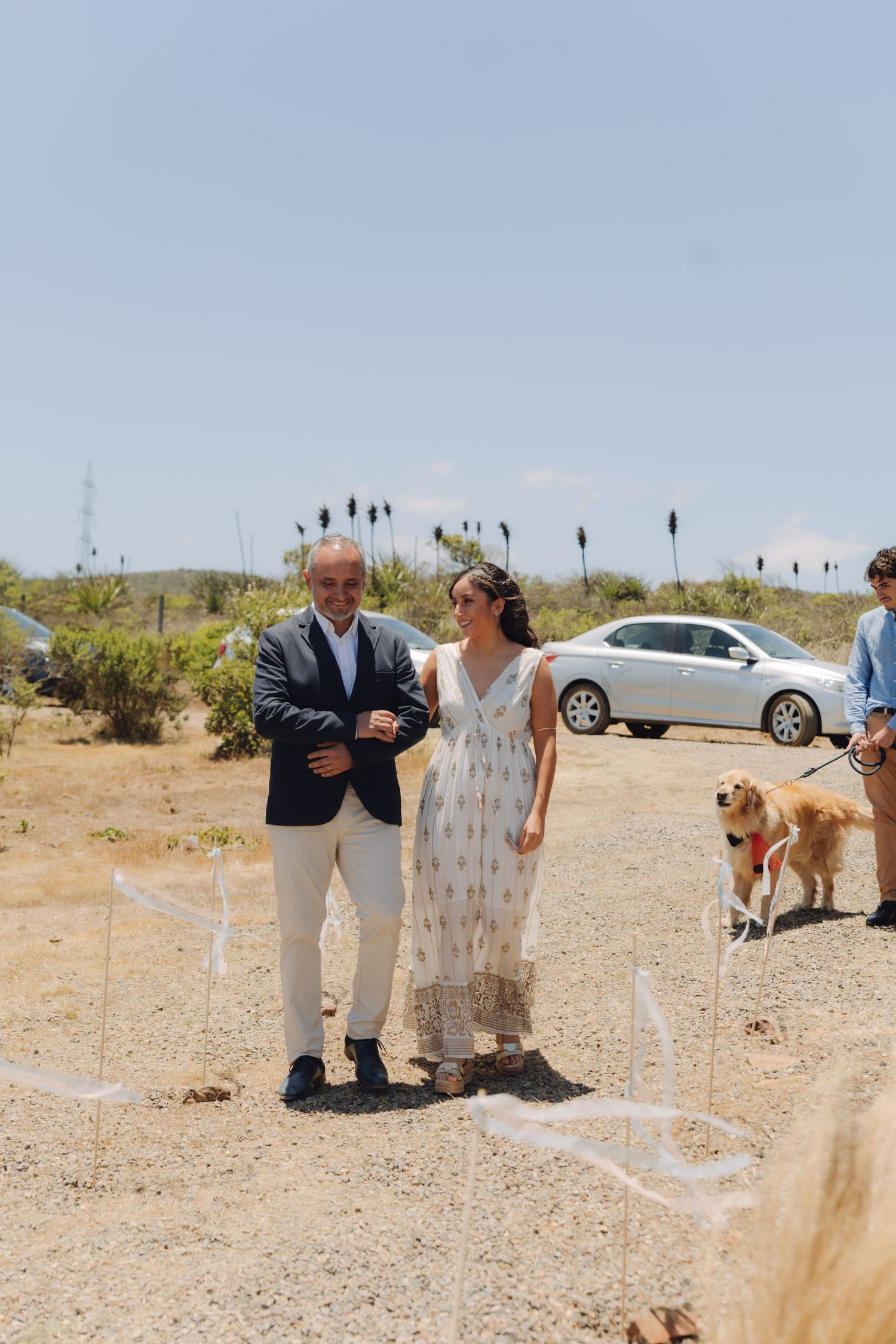 Smiling father escorting bride in white floral dress along sandy outdoor wedding ceremony aisle.