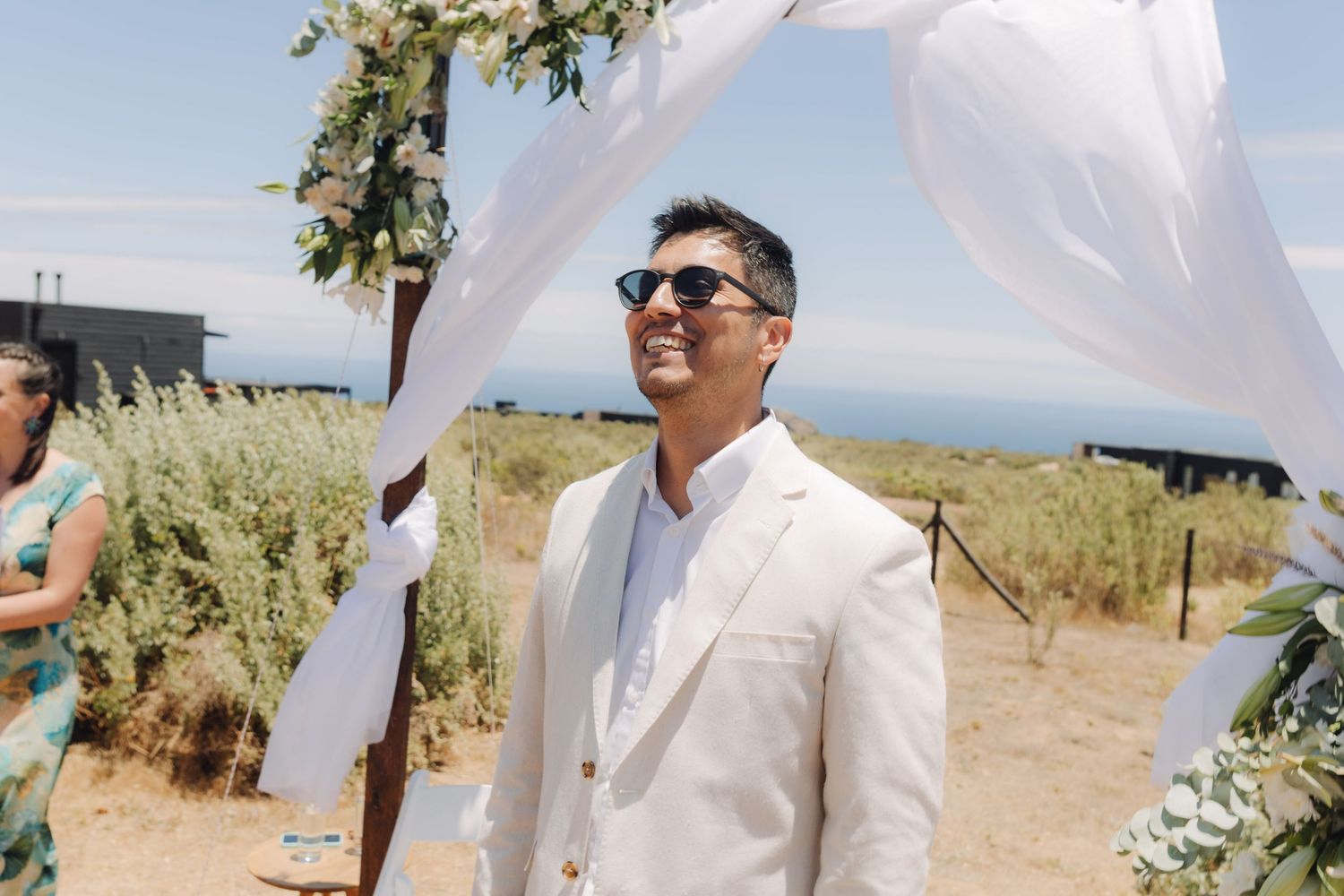 Handsome groom in cream suit smiling under floral wedding arch at sunny outdoor beach ceremony.