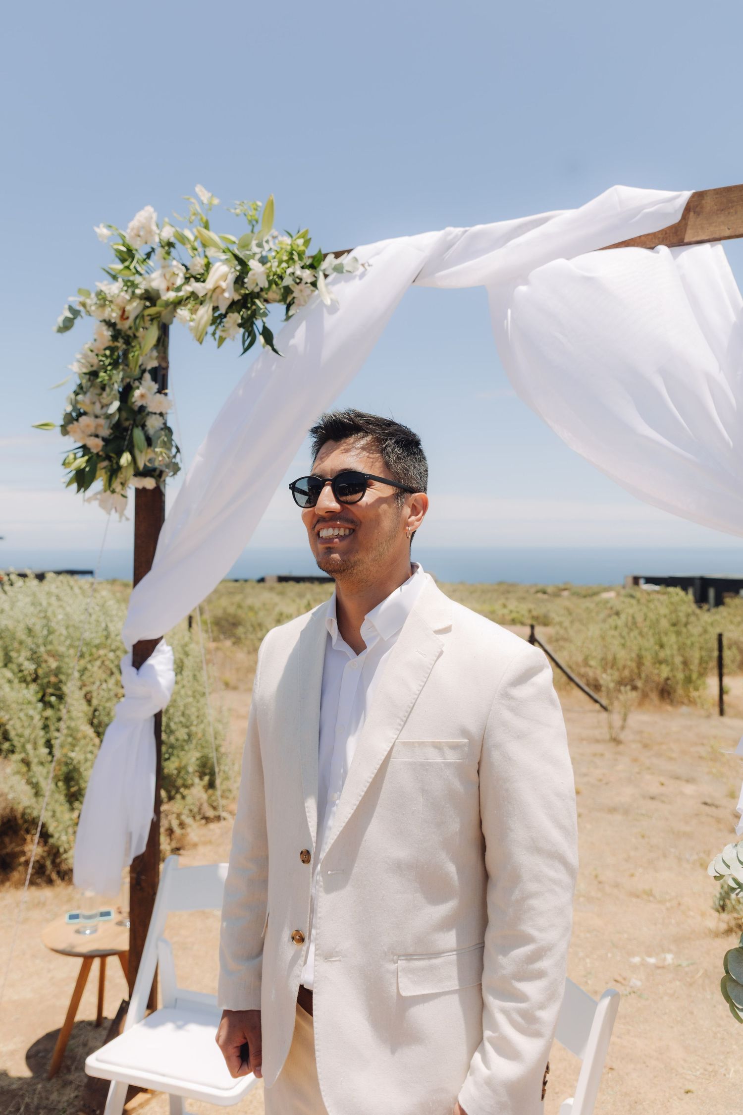 Stylish groom in beige suit and sunglasses standing beneath white draped floral wedding arch outdoors.