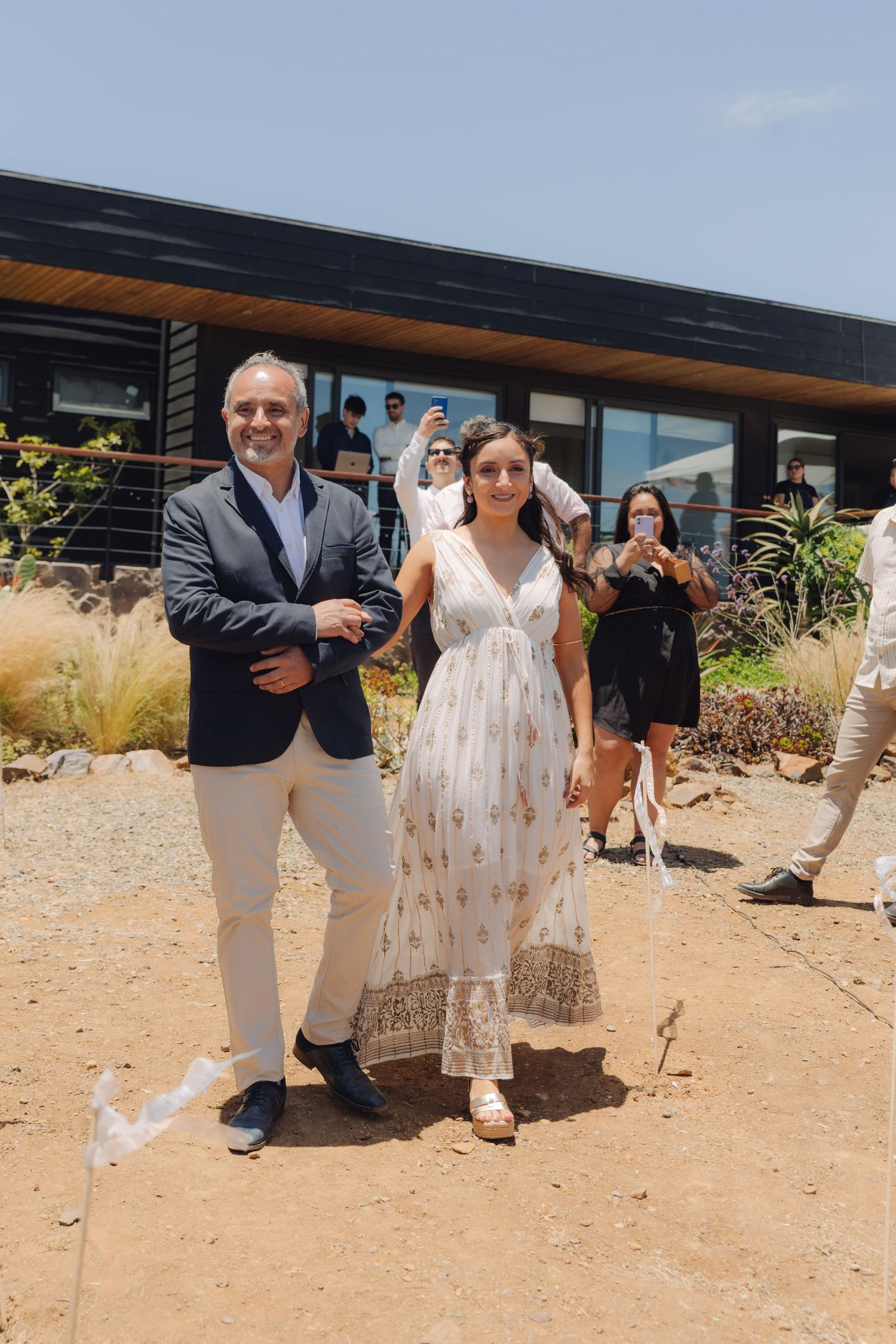 A man in a navy blazer walks a young woman in a white floral dress down an outdoor wedding aisle.
