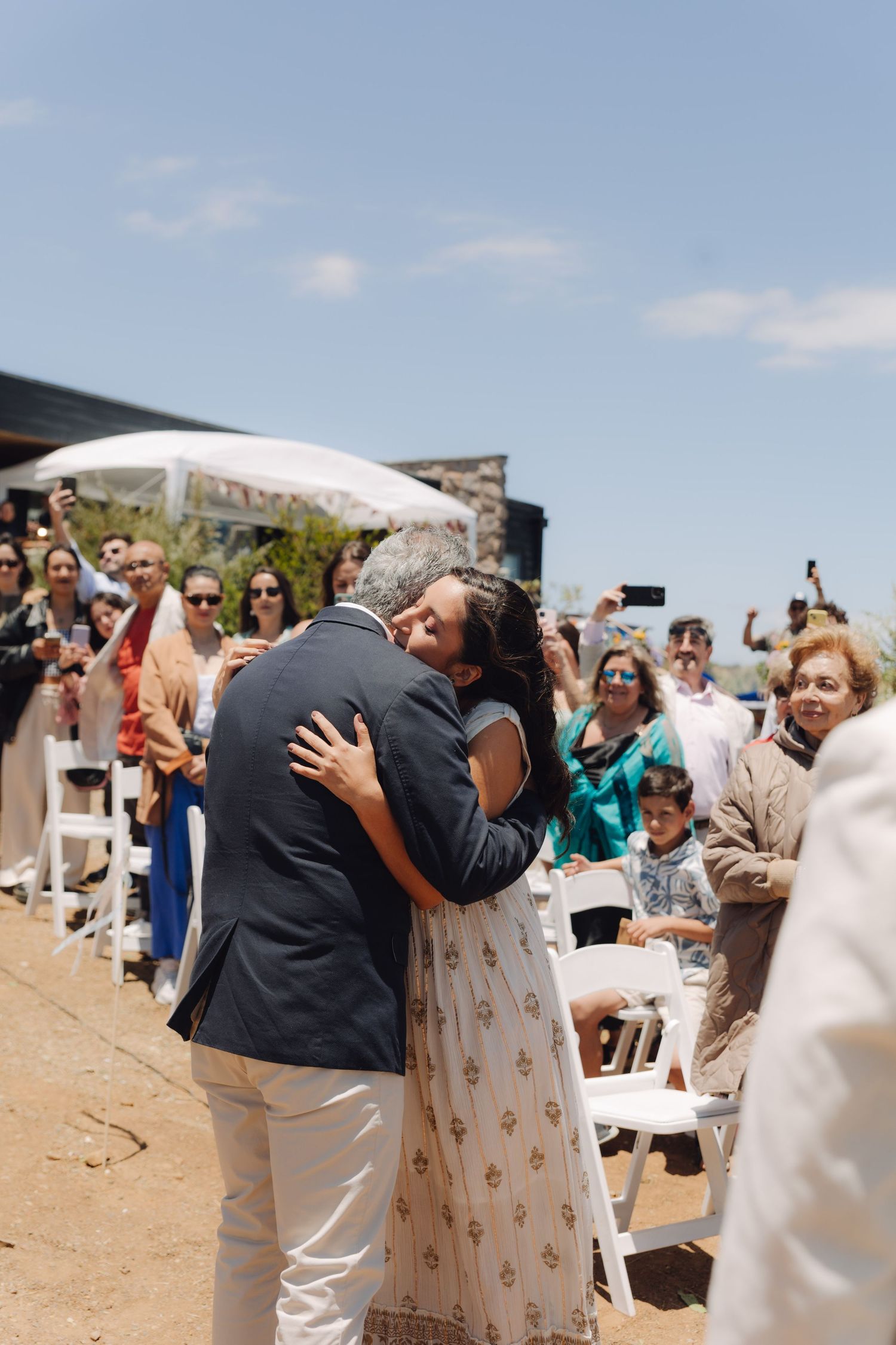 Father of the bride shares an emotional hug with his daughter in a white dress during an outdoor wedding ceremony.