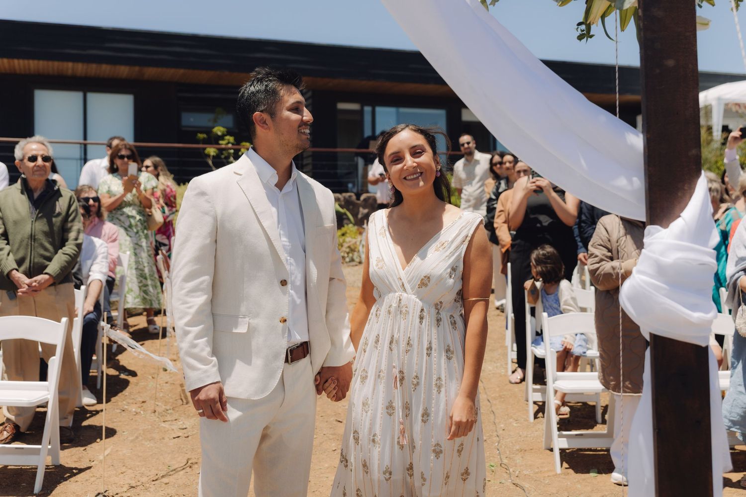 Smiling newlyweds hold hands while walking back down the aisle after their outdoor beach wedding ceremony.