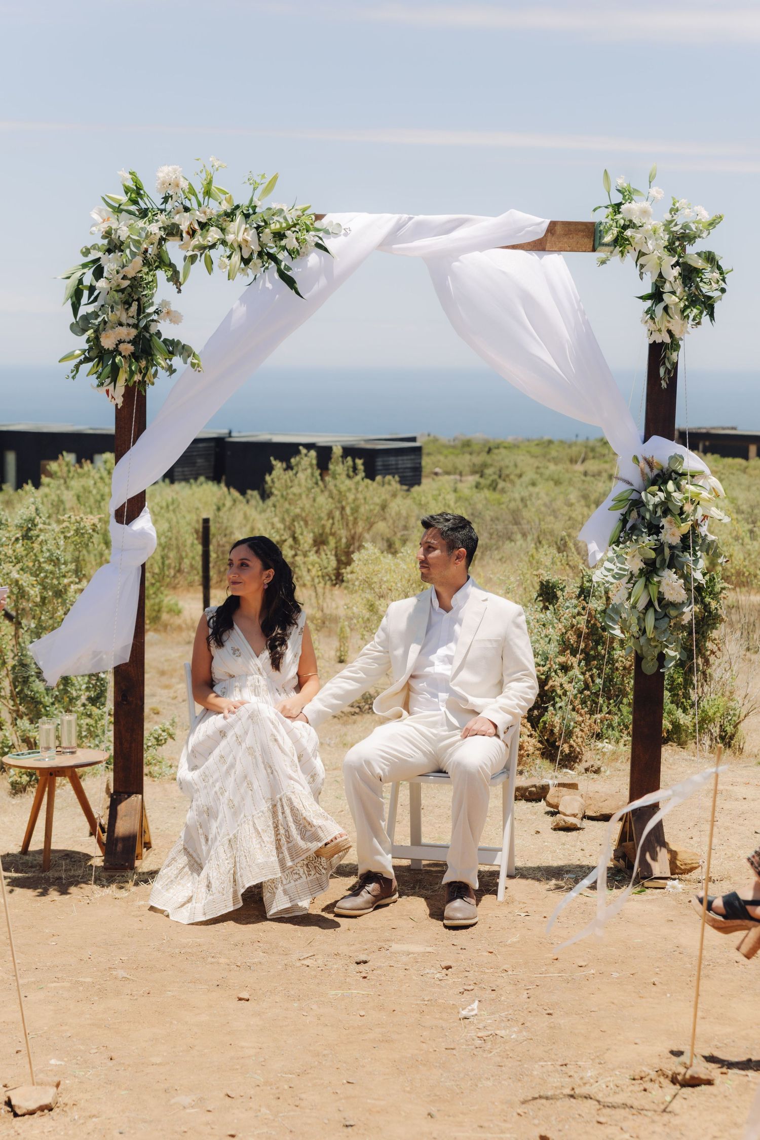 Bride and groom sit together under a white draped floral arch at an outdoor coastal wedding ceremony.