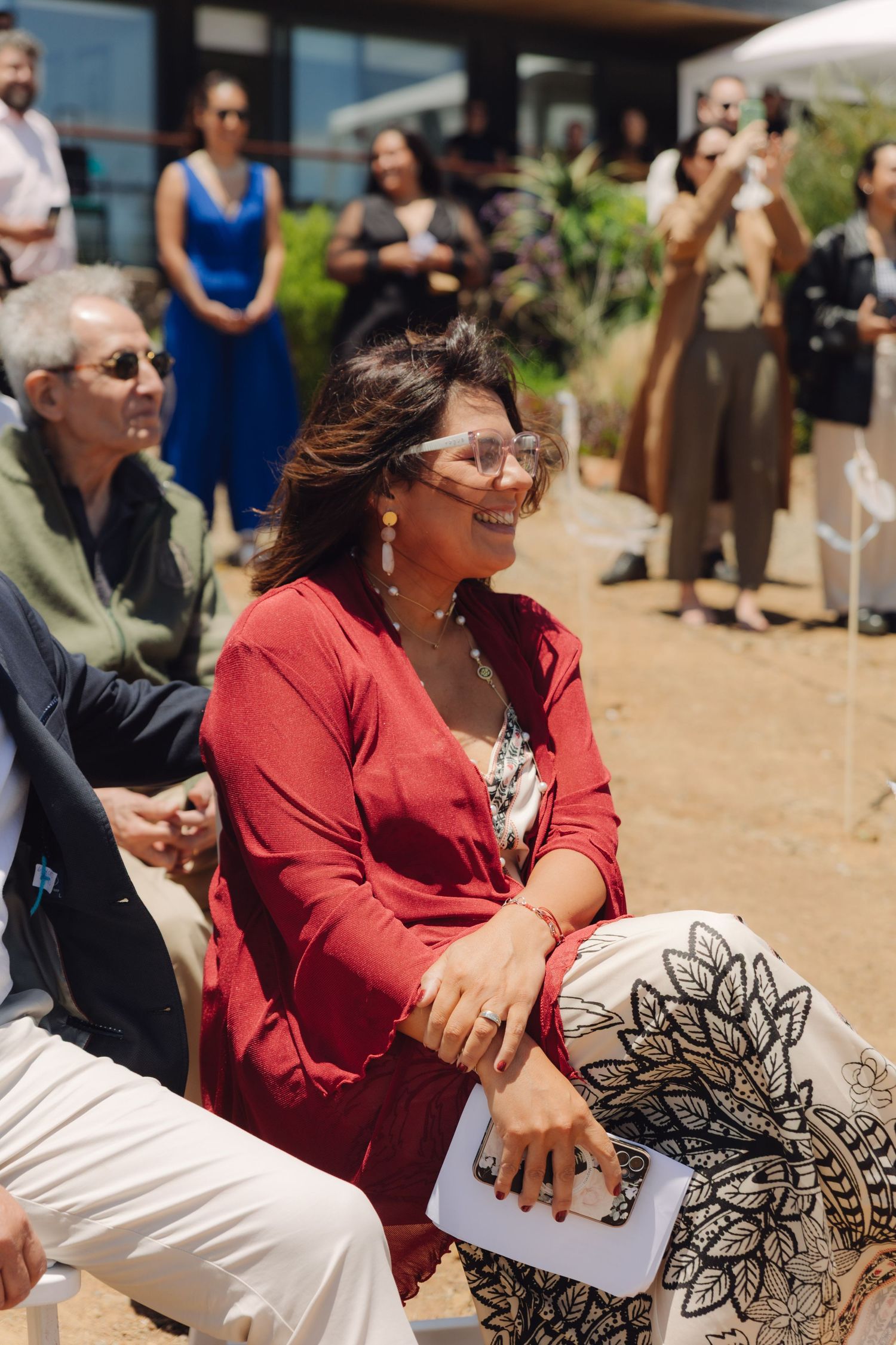 A smiling woman in a red cardigan and patterned skirt enjoys an outdoor wedding ceremony as a guest.