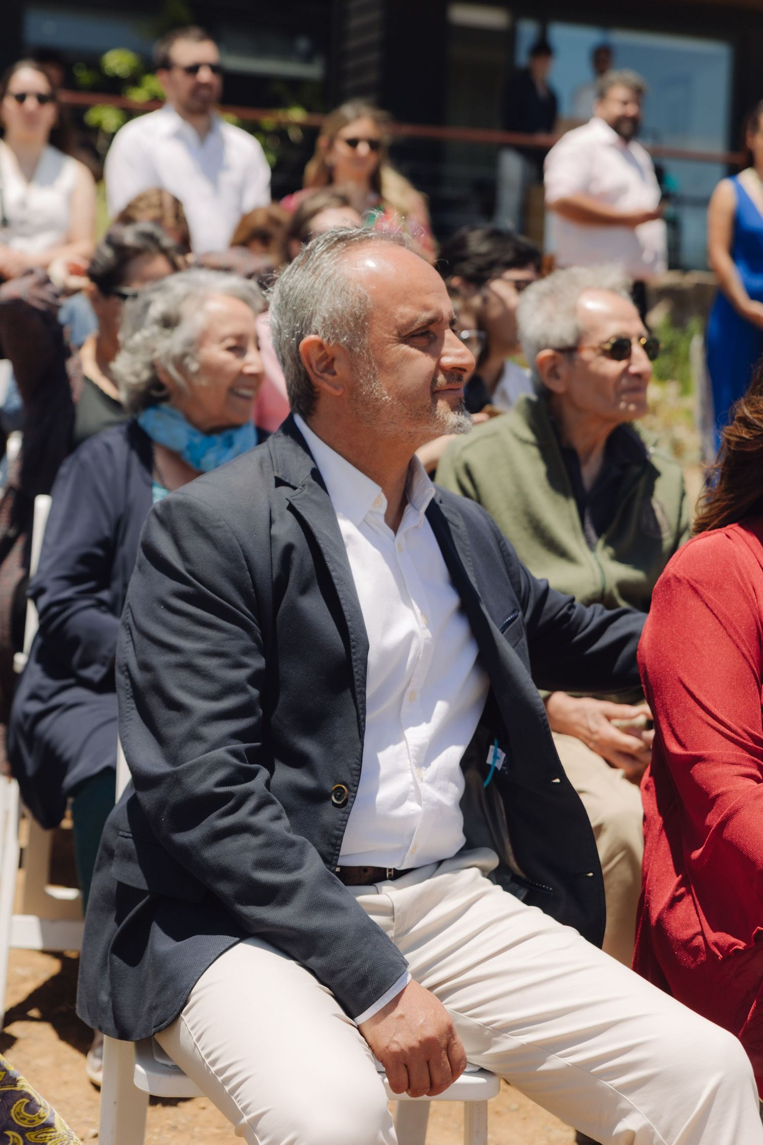 A distinguished man in a navy blazer sits attentively among guests at an outdoor wedding ceremony.