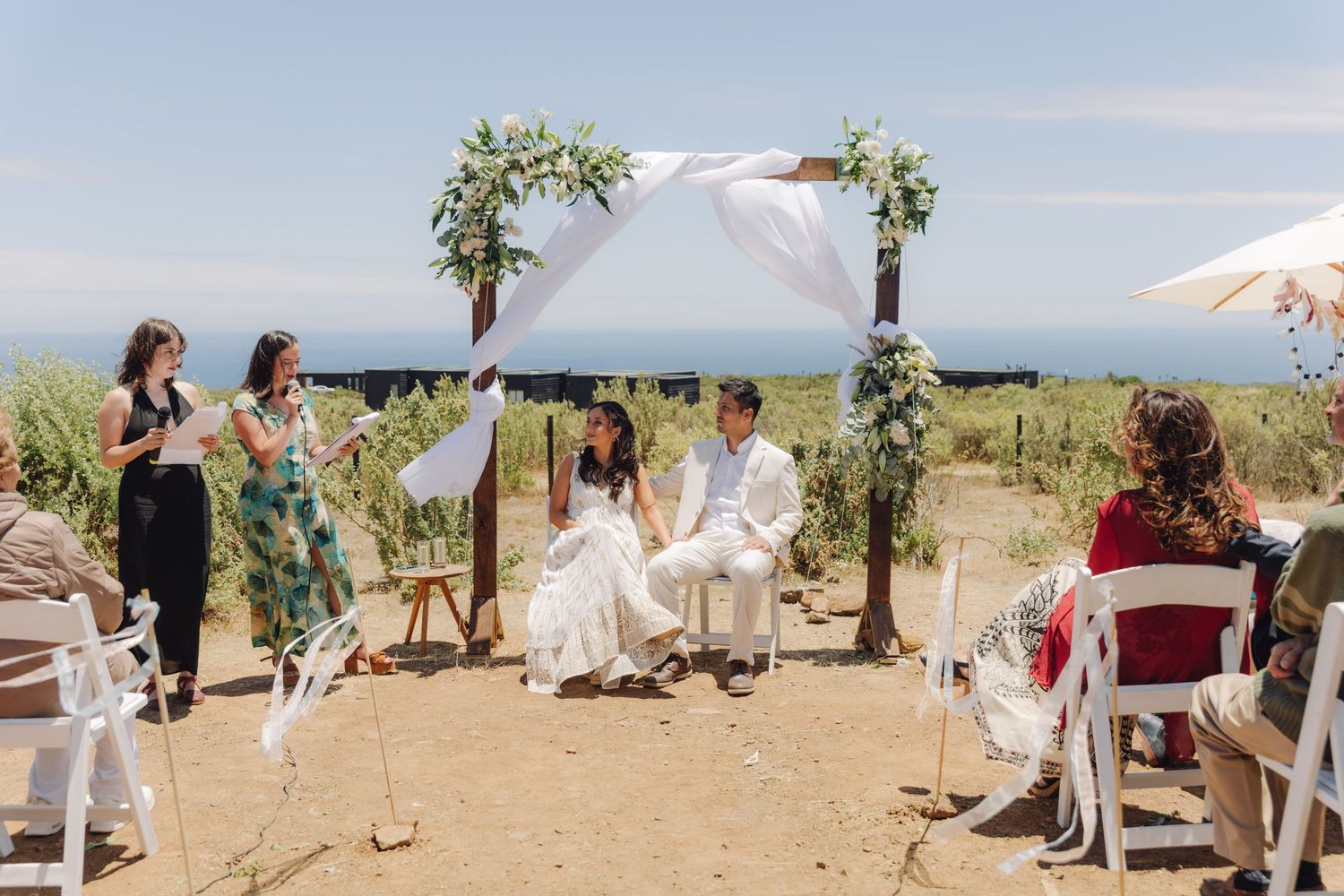 An officiant reads vows as a bride and groom sit under a white draped arch at an outdoor wedding.