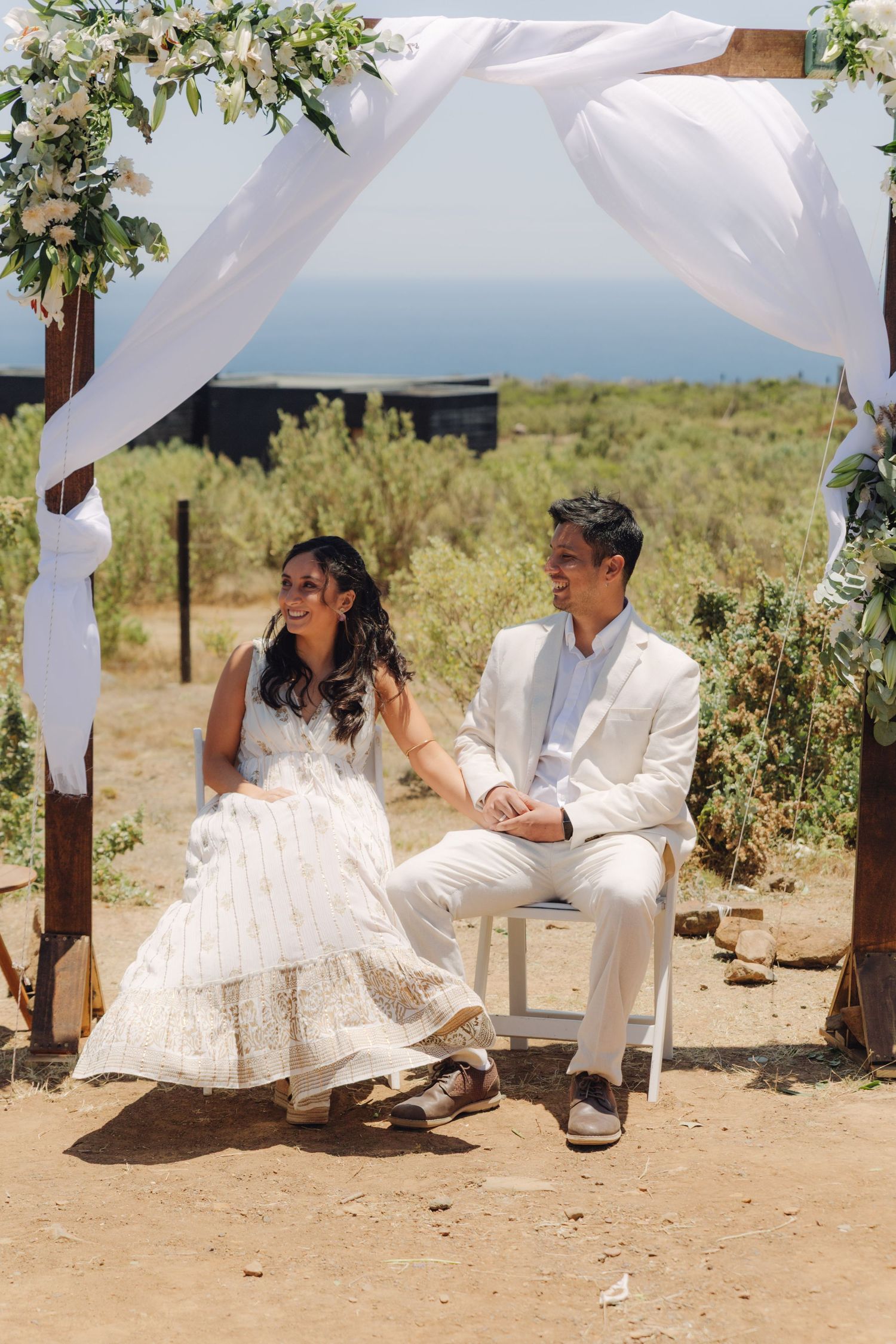 A joyful bride and groom hold hands under a floral decorated arch at a scenic coastal wedding.