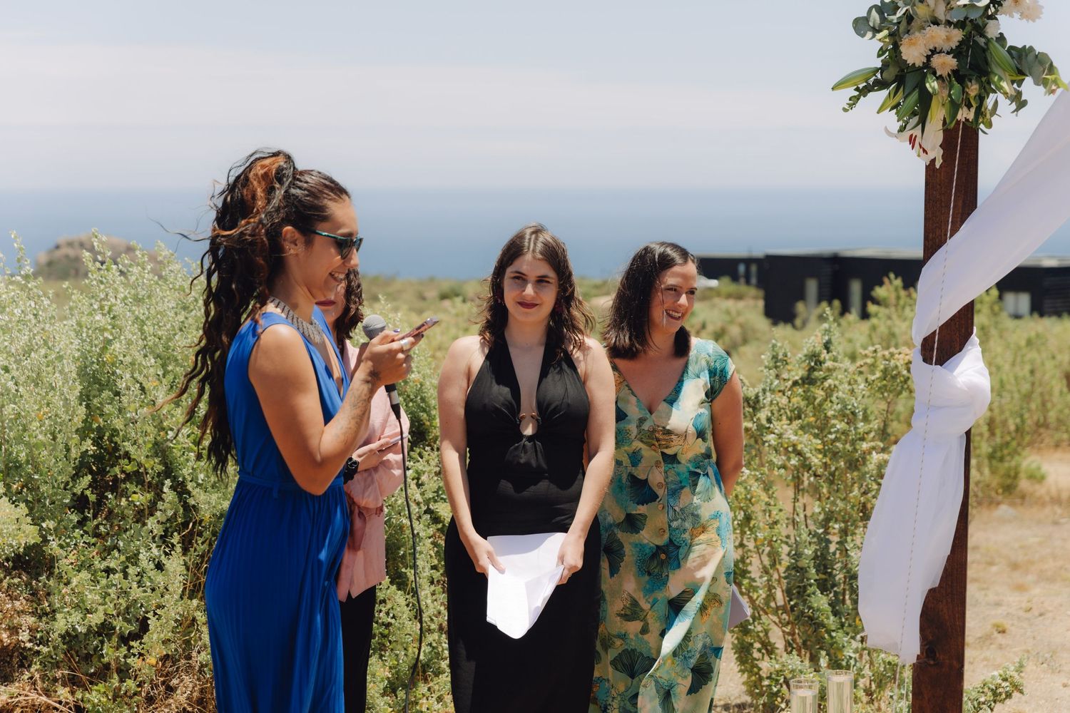 Three women stand near a floral wedding arch, with one reading from a paper at an outdoor ceremony.