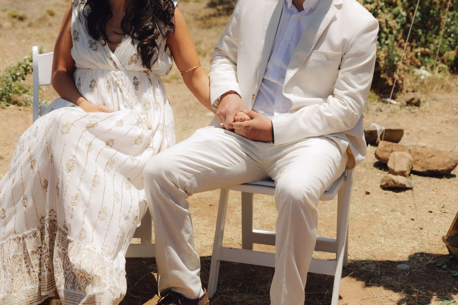 A bride in a layered white dress and groom in a white suit hold hands during their wedding ceremony.