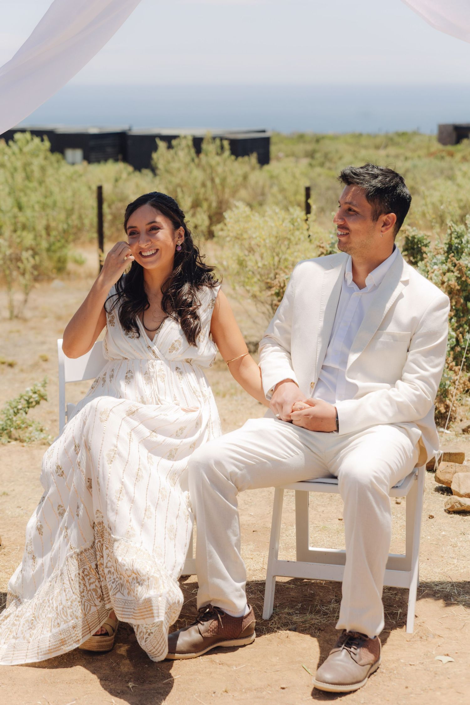 Smiling bride in white lace dress and groom in white suit holding hands at outdoor wedding ceremony.