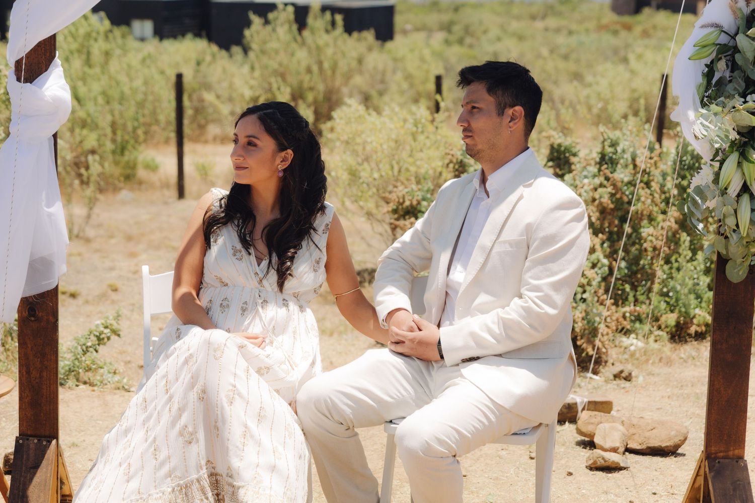 Bride and groom seated together at rustic outdoor wedding ceremony with natural desert landscape.