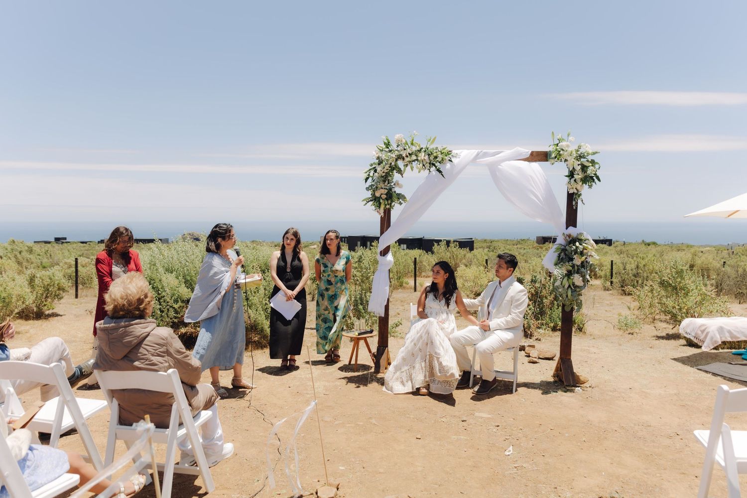 Outdoor wedding ceremony with couple seated under floral arch surrounded by guests in sunny landscape.