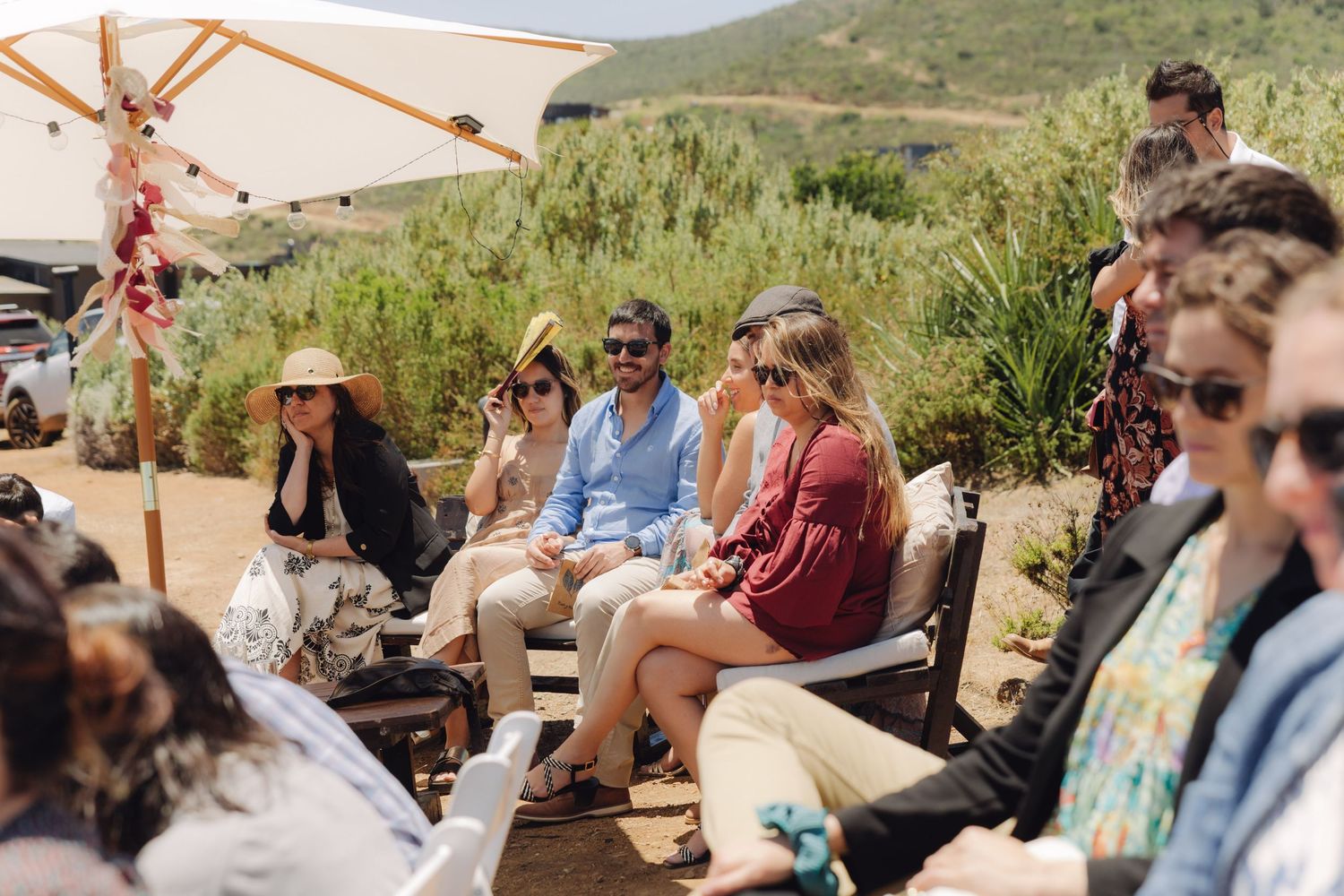 Wedding guests seated outdoors under umbrella enjoying sunny desert ceremony celebration.
