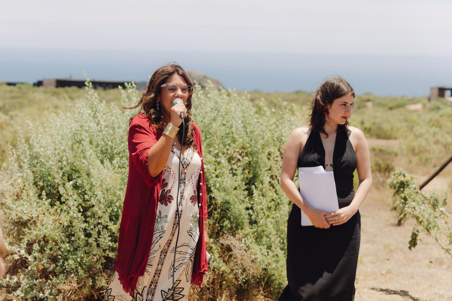 Two women speaking at outdoor wedding ceremony with microphone against scenic coastal backdrop.