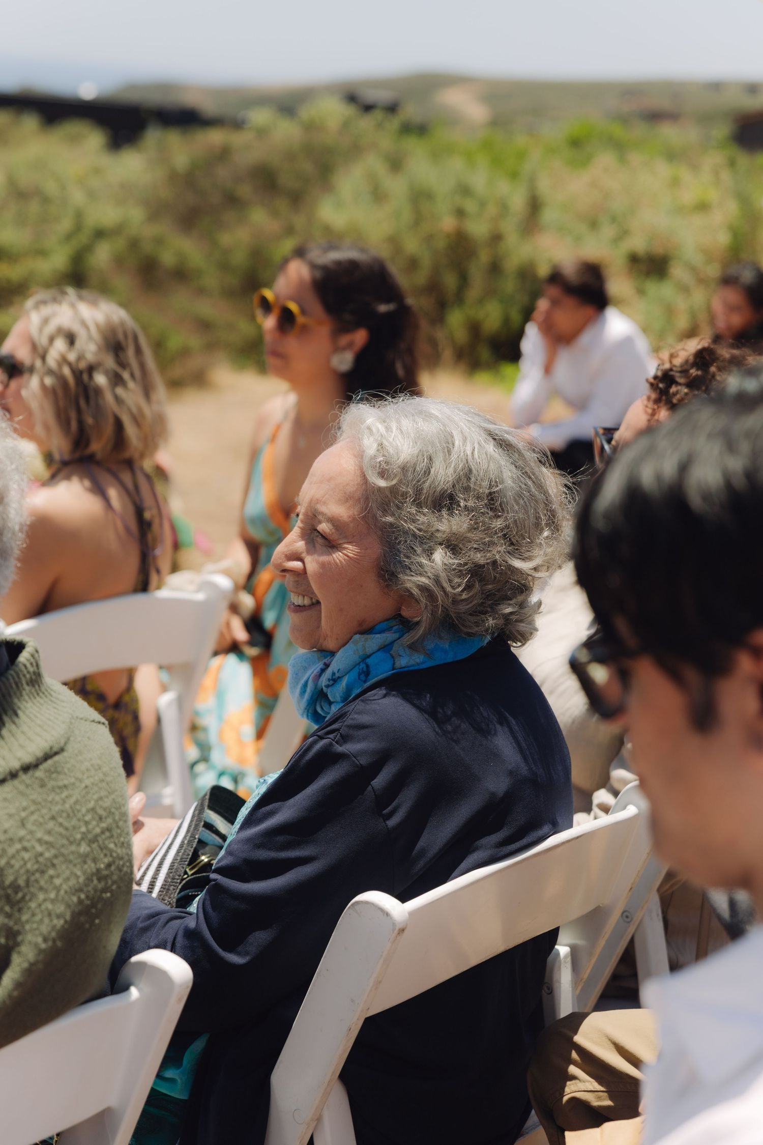 Elderly woman smiling joyfully while seated at outdoor wedding ceremony among other guests.