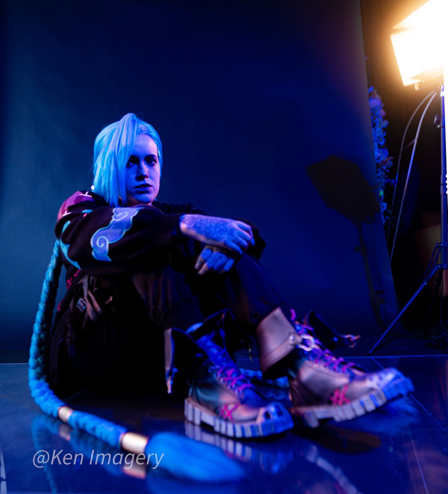 Person with blonde hair sitting on floor in blue-lit studio, wearing black jacket and colorful sneakers, tattoos visible.