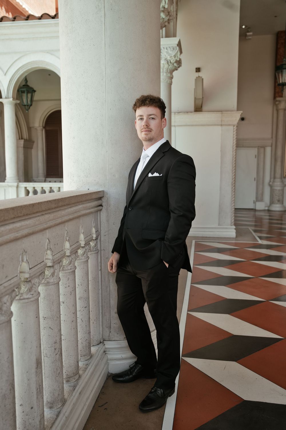 Young man in black suit with white pocket square leaning against marble balustrade in elegant venue.