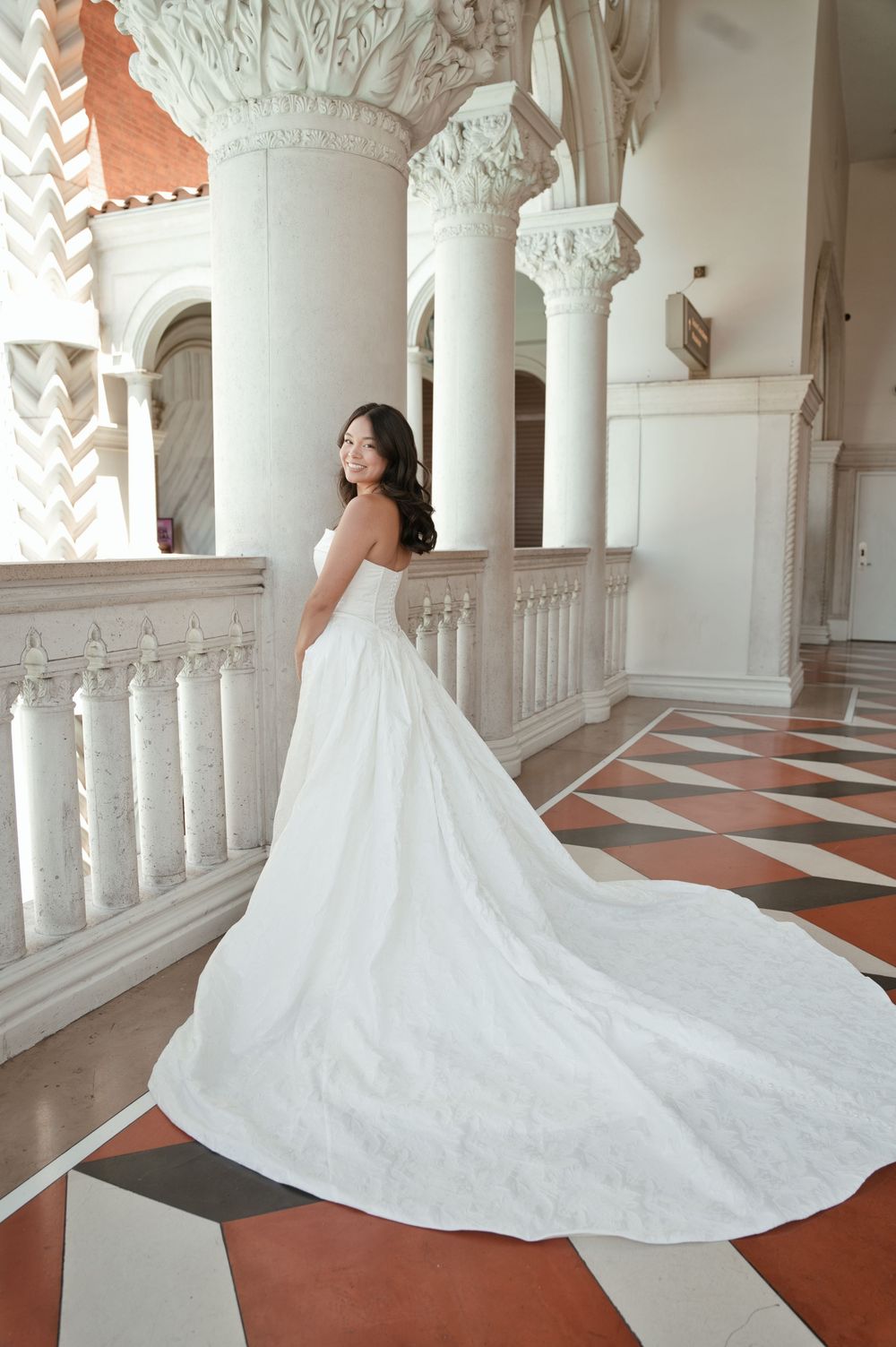 Bride in elegant white strapless ball gown with long train poses in ornate marble hall with decorative columns.
