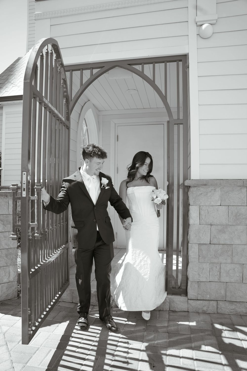Black and white photo of a bride and groom holding hands while exiting a church through ornate iron gates.