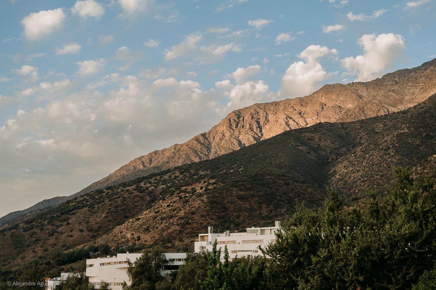 Scenic mountain landscape with lush green slopes, white buildings, and dramatic clouds over a Mediterranean hillside.