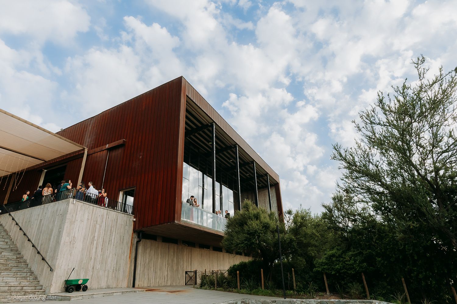 Modern building with rusty red cladding and concrete base, featuring a balcony with guests under a partly cloudy sky.