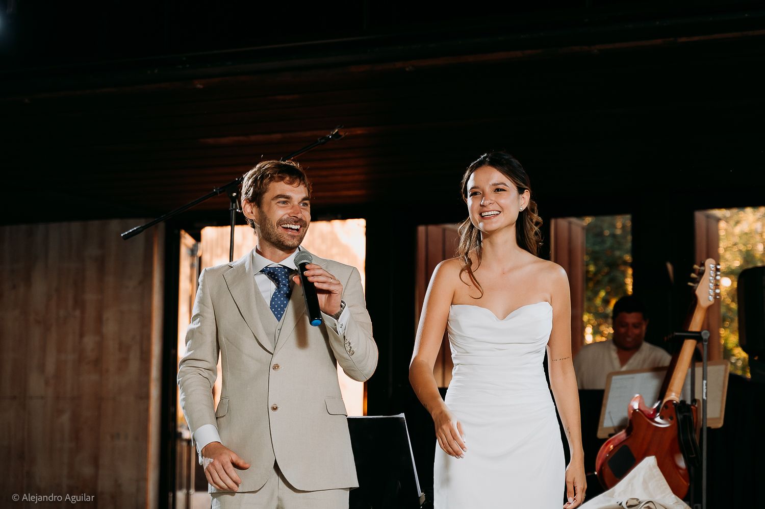 Bride and groom smiling during wedding reception speech, groom in beige suit with blue tie, bride in white strapless gown.