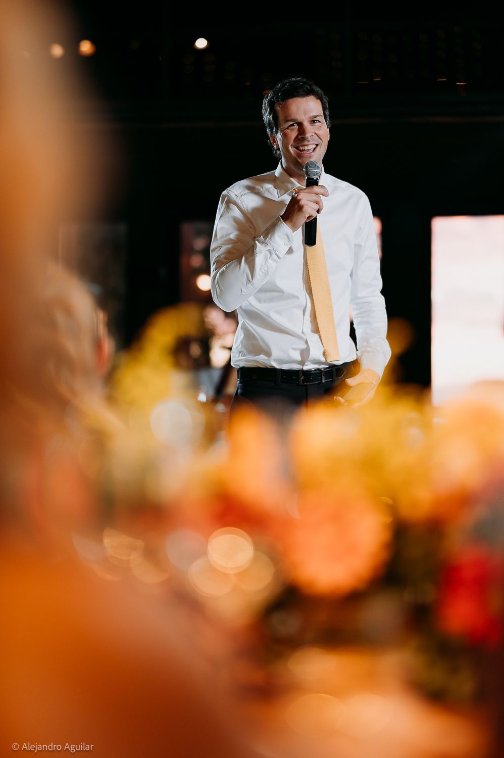 Man in white shirt and gold tie smiling while speaking into microphone at elegant event with floral arrangements.