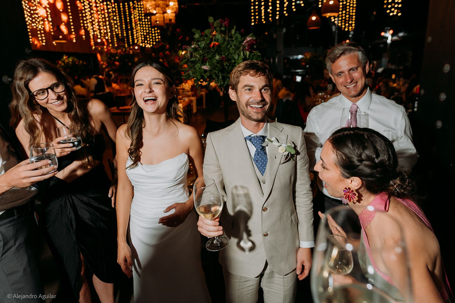 Bride and groom laughing with guests at elegant wedding reception, groom in tan suit holding champagne glass.