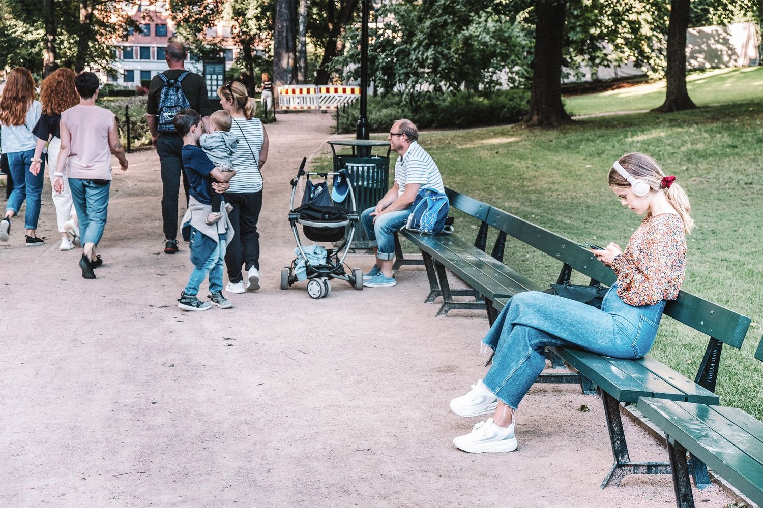 Dívka se sluchátky na lavičce mezi lidmi v městském parku.