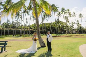 Bride and groom stand beneath tall palm trees on lush green lawn at tropical outdoor wedding ceremony.