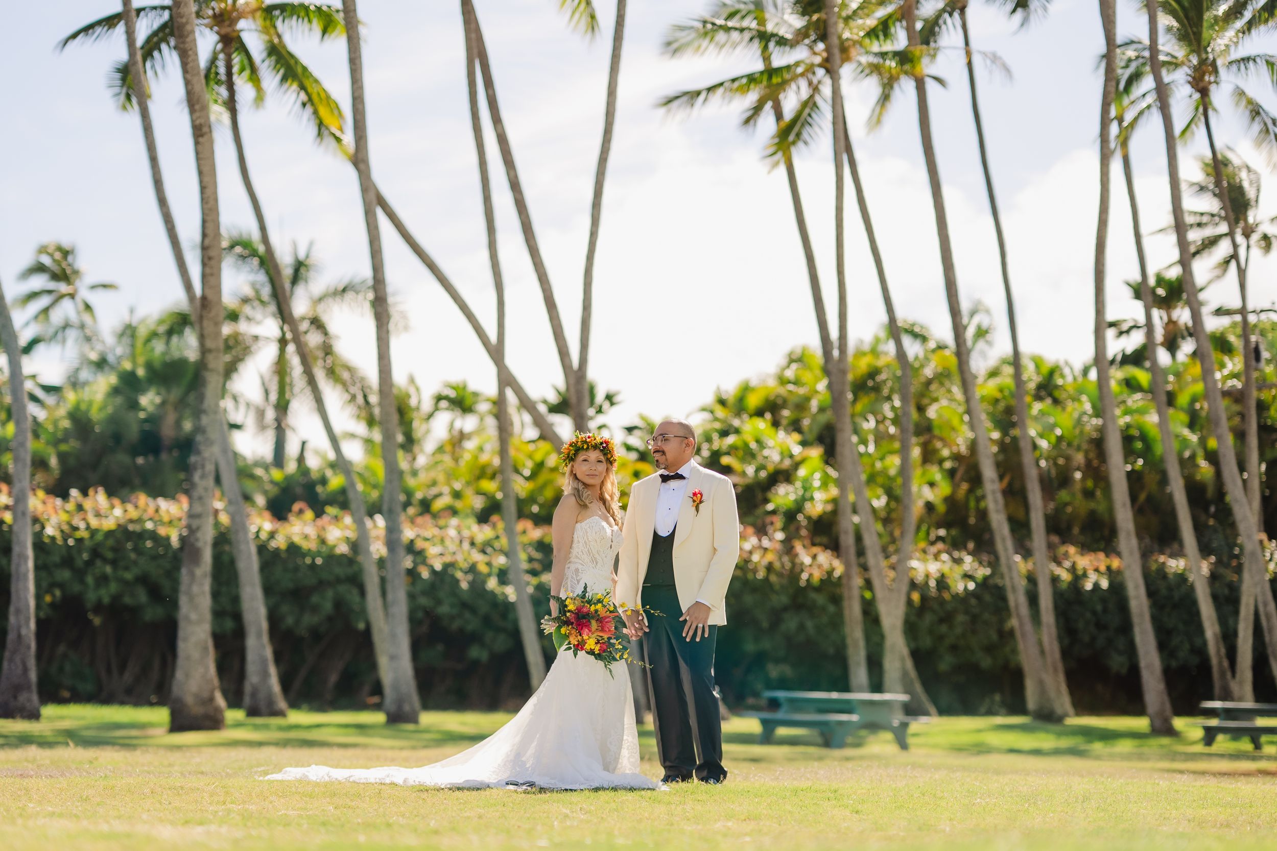 Bride and groom pose on tropical lawn surrounded by tall palm trees on their Hawaiian wedding day.