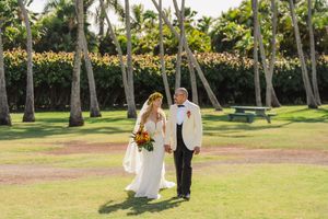 Bride and groom walking hand in hand on lush green lawn surrounded by tropical palm trees at outdoor Hawaii wedding.