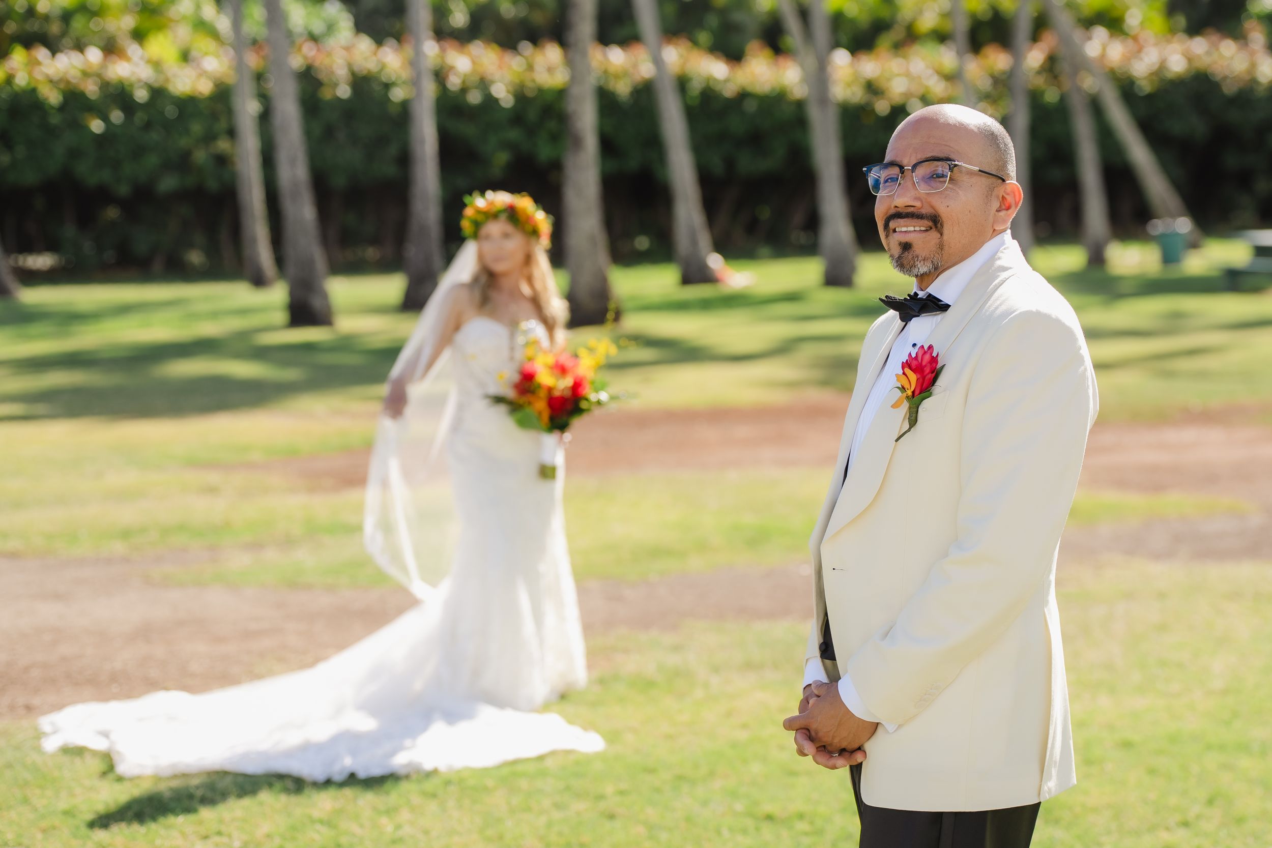 Groom in white tuxedo smiles as bride in floral crown holds bouquet during outdoor Hawaii wedding first look.