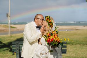 Bride and groom share a kiss with a rainbow over the ocean behind them, bride holding vibrant tropical bouquet.