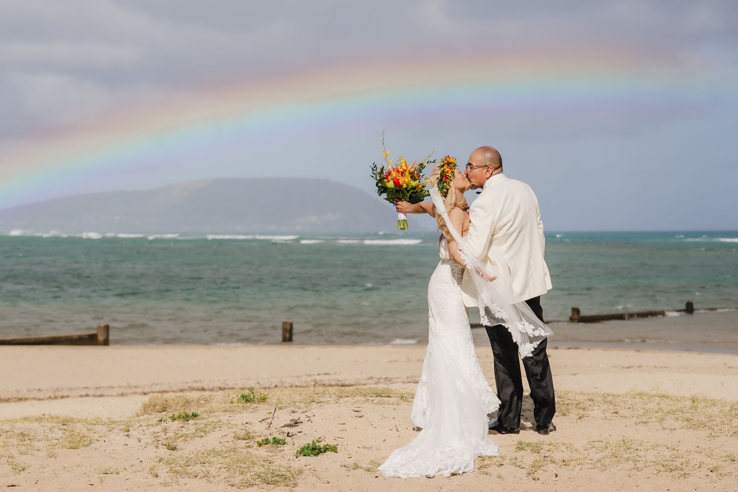 Bride and groom kissing on a beach with a rainbow over the ocean, bride holding a tropical bouquet.