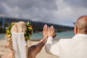 Newlywed couple on beach holding up hands showing wedding rings, bride wearing floral crown and veil.