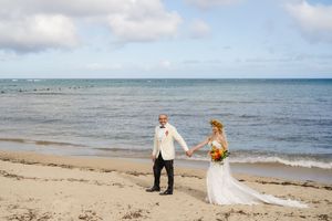 Bride and groom hold hands on a sandy beach with calm blue ocean waves and partly cloudy skies behind them.