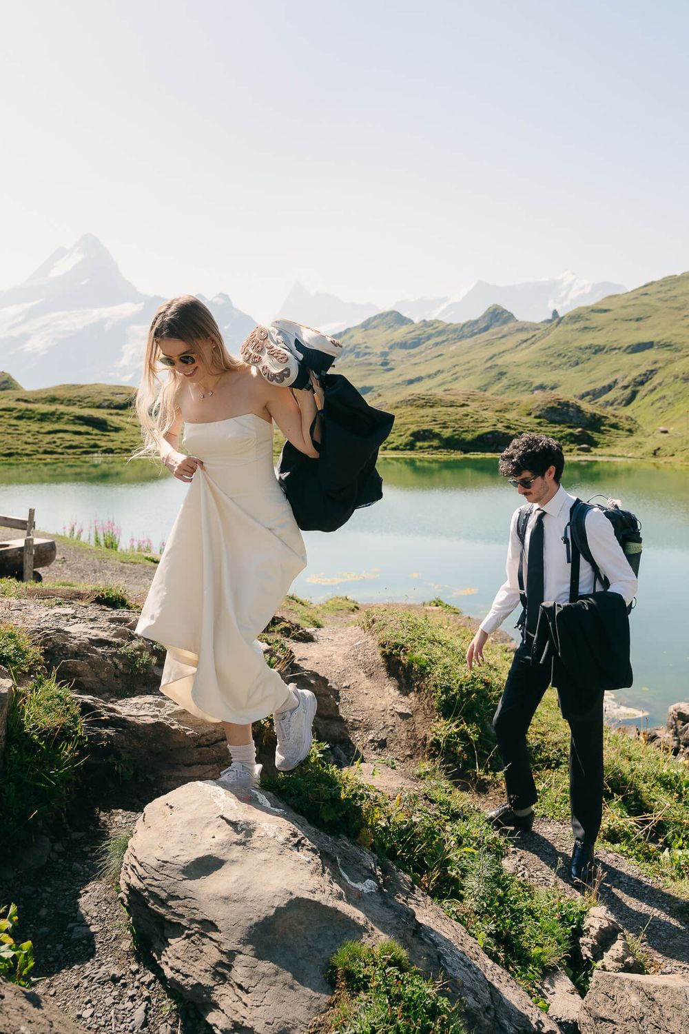 Bride in white dress and groom in black suit hiking rocky alpine trail beside turquoise mountain lake in Switzerland.