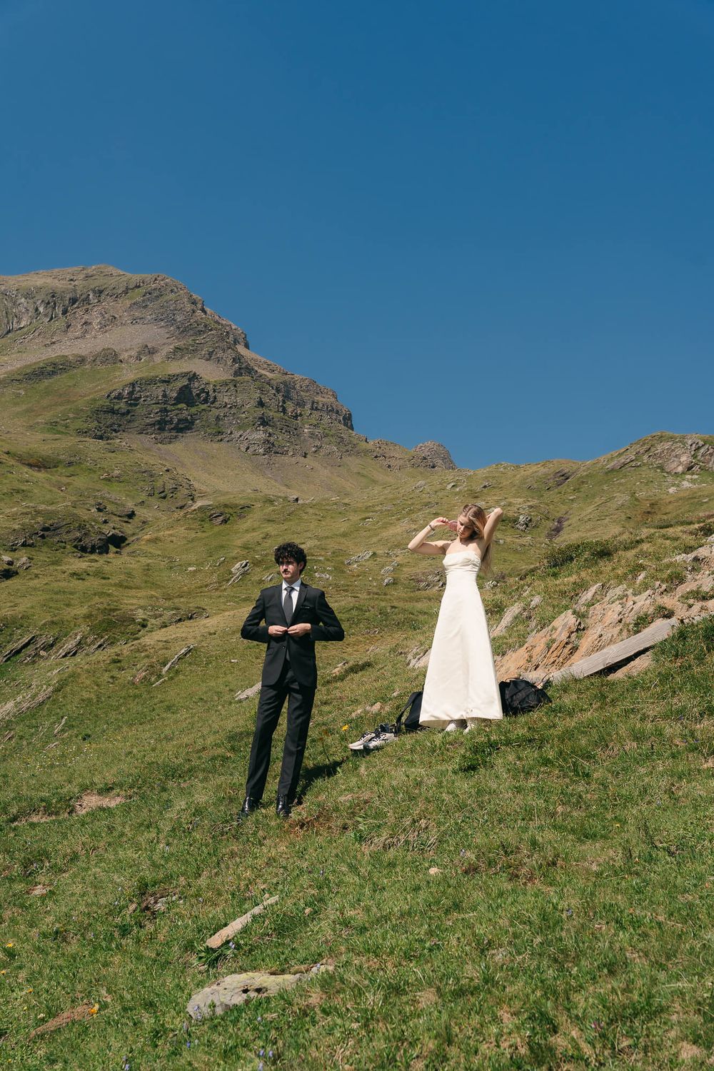 Bride in white dress and groom in black suit pose on green mountain hillside under clear blue sky.
