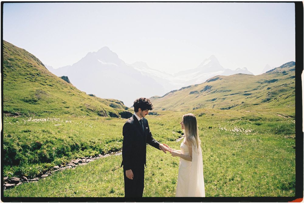 Bride and groom hold hands in lush green alpine meadow with misty mountain peaks in background.