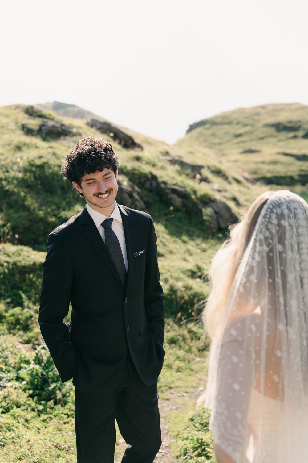 Groom in black suit smiling at bride wearing dotted veil, with lush green hills in background at outdoor wedding.