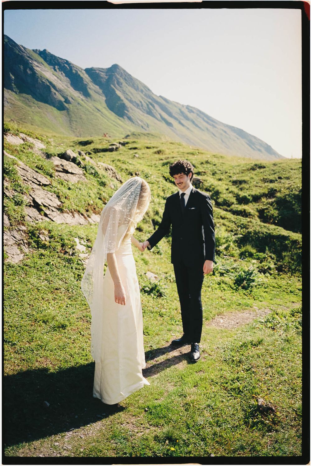 Bride in white gown and veil with groom in black suit on green mountain path on sunny wedding day.