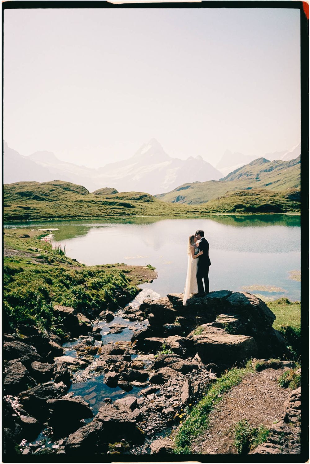 A couple shares a romantic embrace on rocks beside a serene alpine lake with snow-capped mountains in the background.