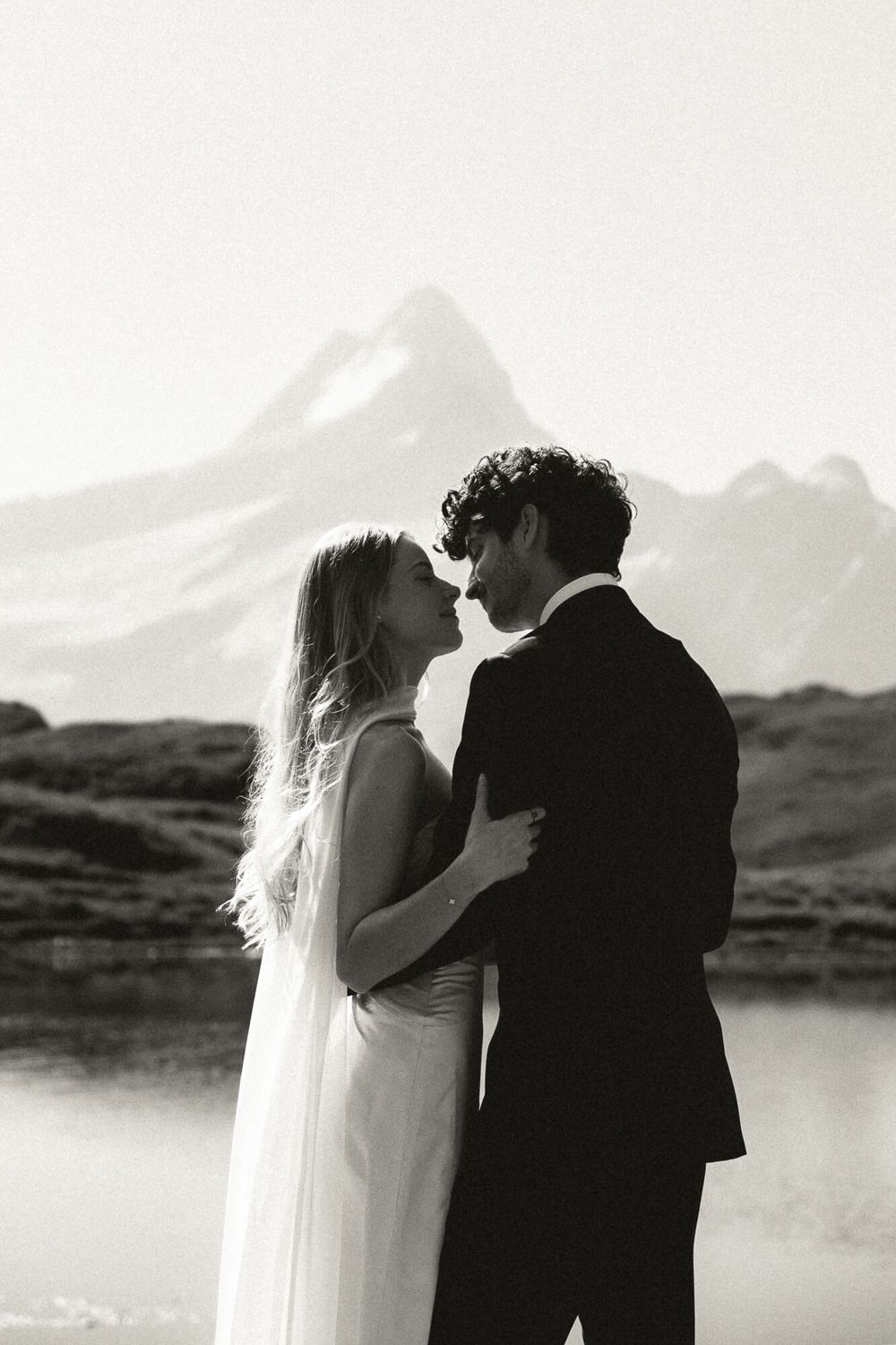 Black and white photo of a couple in wedding attire almost kissing with a mountain lake backdrop.