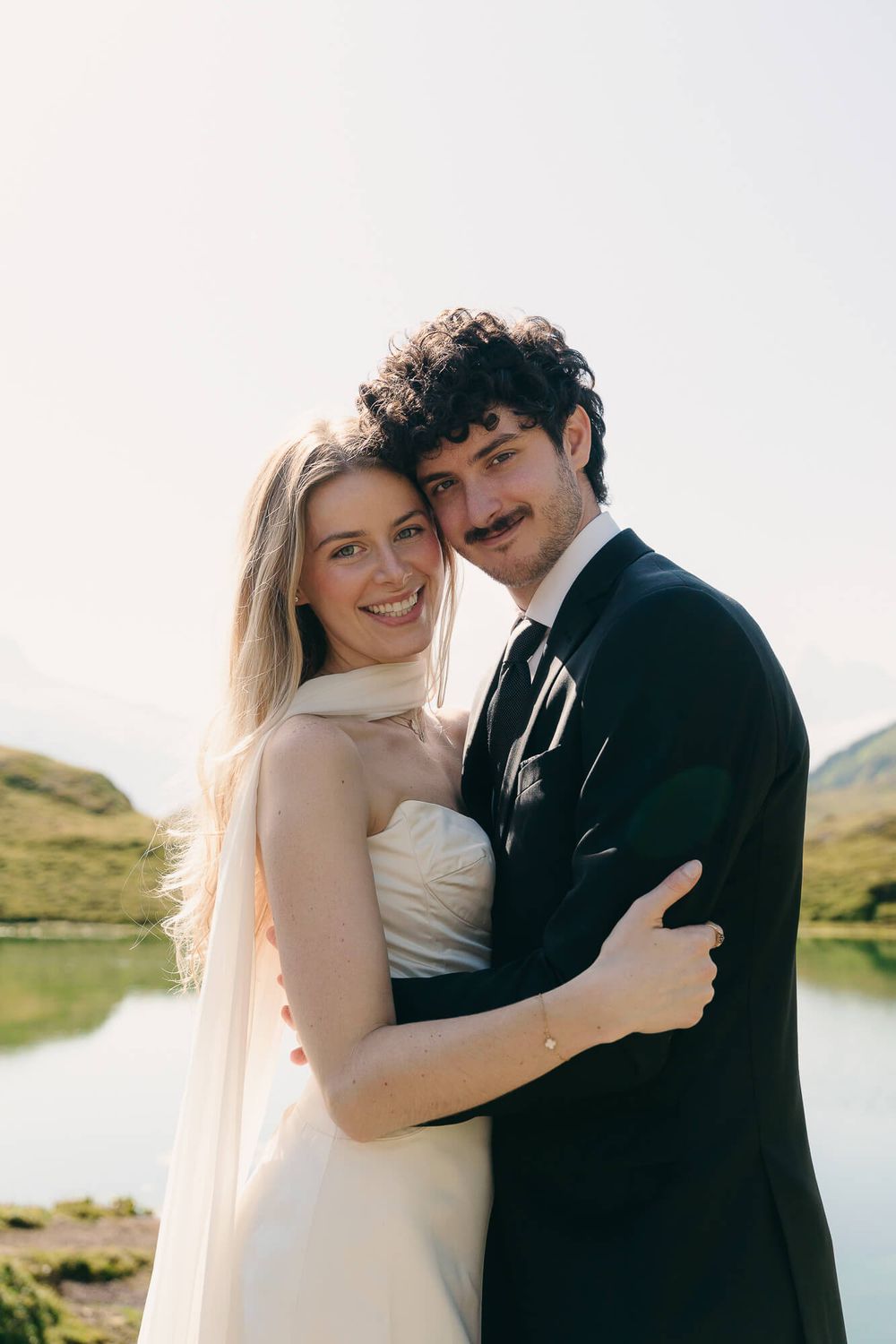 A smiling bride and groom embrace outdoors near a scenic lake, wearing a white dress and dark suit with tie.