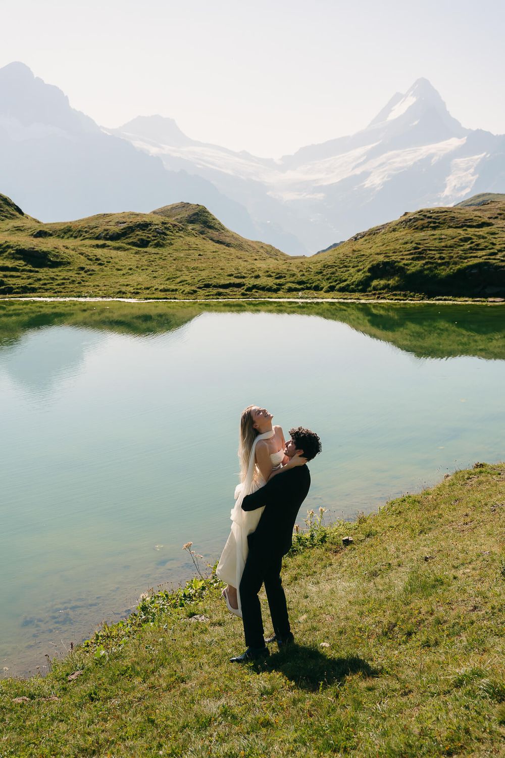 Couple embracing by a serene alpine lake with snow-capped mountains reflected in the water during their wedding day.