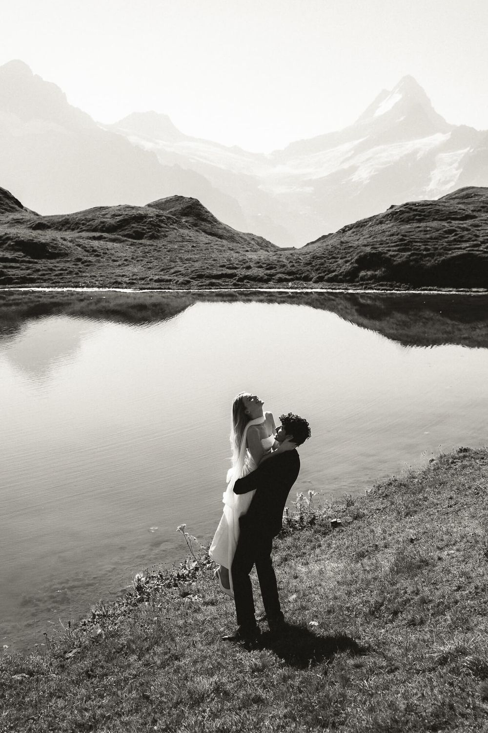 Black and white photo of a couple embracing by a serene mountain lake with snow-capped peaks reflecting in the water.