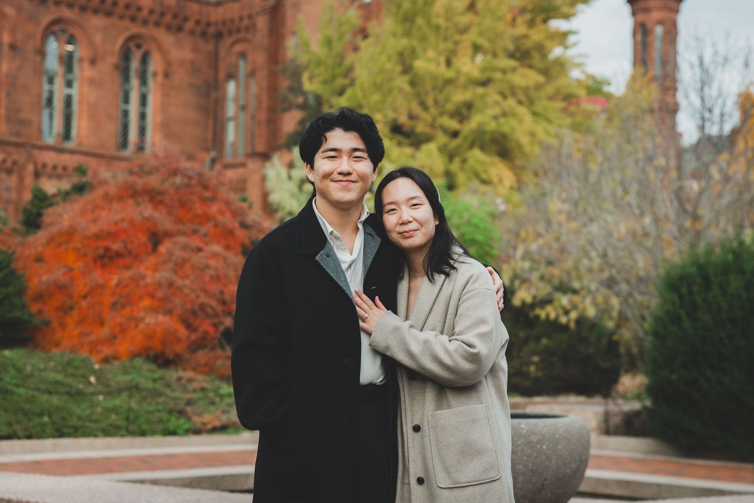Couple smiling together outdoors in front of a red brick building with colorful autumn foliage.